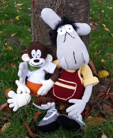 Zwei gehäkelte Amigurumi-Figuren: brauner Affe mit orangefarbener Hose und graue Figur mit Latzhose, sitzend im Gras vor einem Baum