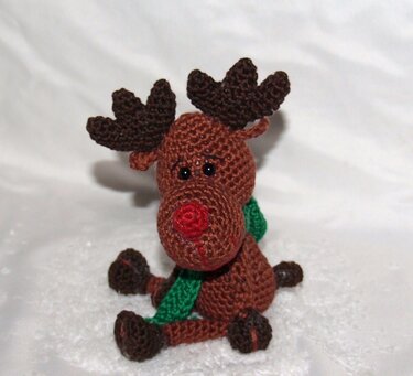 Gehäkeltes Amigurumi-Rentier in Braun mit roter Nase, dunkelbraunen Geweihen und grünem Schal, sitzend