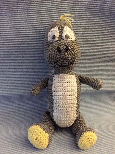 Sitzender, gehäkelter grauer Dinosaurier mit weißem Bauch und gelben Akzenten vor gestreiftem Stoff