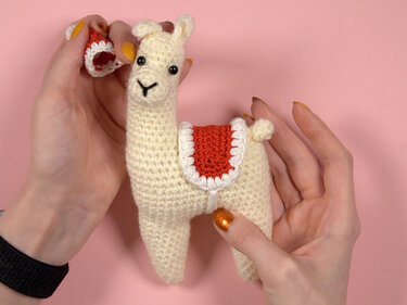Cremefarbenes gehäkeltes Alpaka-Amigurumi mit rotem Sattel und Santa-Mütze, in Händen gehalten vor rosa Hintergrund