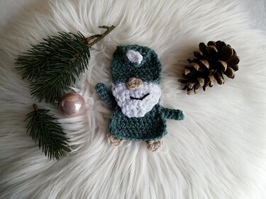 Gehäkelter grüner Wichtel mit weißem Bart, umgeben von Tannenzweigen, Weihnachtskugel und Tannenzapfen auf weißem Fell