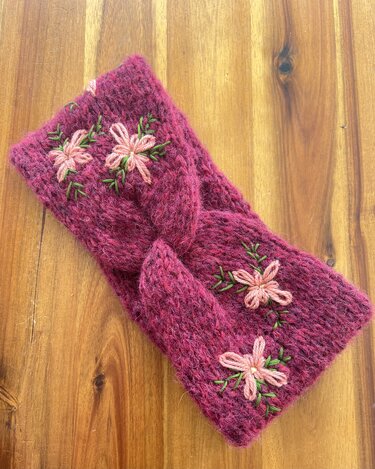 Himbeerfarbenes gestricktes Stirnband mit rosa Blütenstickerei und grünen Blättern auf Holzhintergrund
