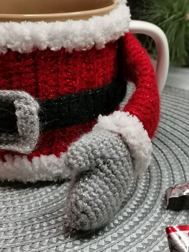 Nahaufnahme eines gehäkelten roten Nikolaus-Tassenwärmers mit weißem Flauschrand, schwarzem Gürtel, grauer Schnalle und grauem Stiefel an einer Tasse auf grauem Untergrund.