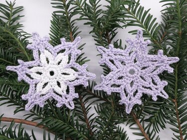 Zwei gehäkelte Schneeflocken in Lavendel und Weiß auf Tannenzweigen