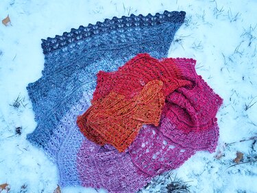 Drei gehäkelte Tücher in leuchtenden Blau-, Rot- und Pinktönen auf Schneehintergrund.