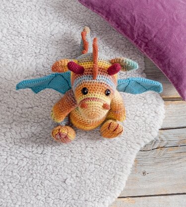Gehäkelter Amigurumi‑Drache in Regenbogenfarben mit blauen Flügeln auf hellem Teppich