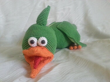 Gehäkelter grüner Amigurumi-Dino mit orangefarbenem Maul, roter Zunge und großen weißen Augen auf hellem Stoff.