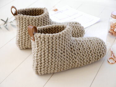 Pair of beige chunky knit ankle slippers with leather pull tabs on a light wooden floor