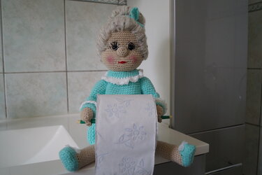 Gehäkelte Oma-Puppe mit grauem Haar und türkisfarbenem Kleid hält eine Toilettenpapierrolle auf einer Ablage.
