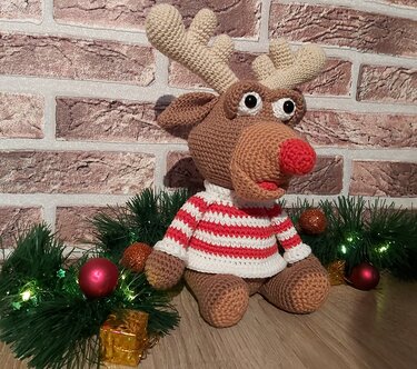 Sitzendes Rentier-Amigurumi mit roter Nase und rot-weiß gestreiftem Pullover zwischen Tannengrün, Kugeln und Geschenkpäckchen.