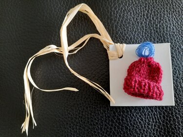 Rote gestrickte Mini-Mütze mit blauem Bommel auf weißer Karte mit Bastband.
