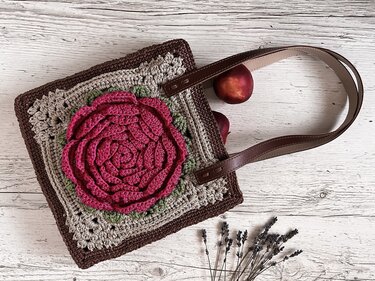 Square crochet shoulder bag with a large pink crocheted rose, brown straps, and two peaches visible nearby on whitewashed wood.