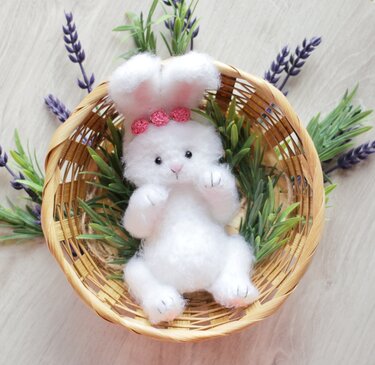 Close-up of small white amigurumi rabbit with fuzzy texture, stitched paws and pink flower crown in a woven basket with lavender accents