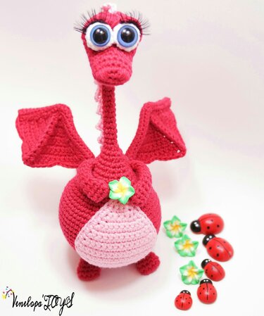 Red crocheted amigurumi dragon with large round eyes and a small flower on its chest