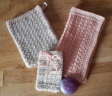 Zwei gehäkelte Tücher in Beige und Rosa, dazu ein gehäkelter Seifenbeutel mit Band und eine lila Seife auf einem Holztisch.