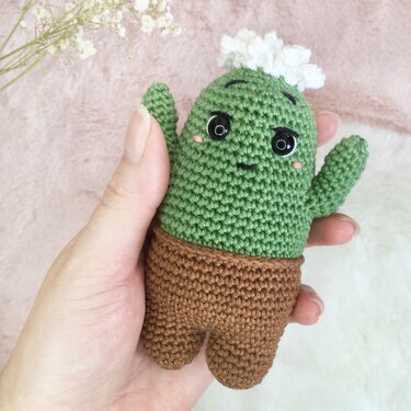 Hand hält gehäkelten grünen Kaktus-Amigurumi im braunen Topf mit schwarzen Augen, rosa Wangen und weißer Blüte.