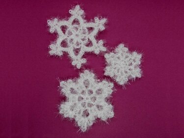 Drei weiße, gehäkelte Schneeflocken aus flauschigem Effektgarn auf magentafarbenem Hintergrund