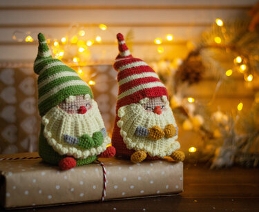 Green and red striped crochet gnomes with long beards sitting on a wrapped gift with fairy lights in the background.