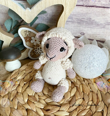 Gehäkeltes Amigurumi-Schaf in Beige und Creme, sitzend auf geflochtener Unterlage mit Holzdeko und kleinem Pilz