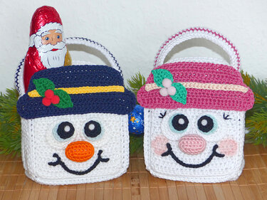Zwei weiße, gehäkelte Körbe mit aufgenähten Schneemann-Gesichtern, einer mit blauer Mütze und Stechpalmen-Applikation, einer mit rosa Mütze und Beeren-Applikation, jeweils mit Henkeln.