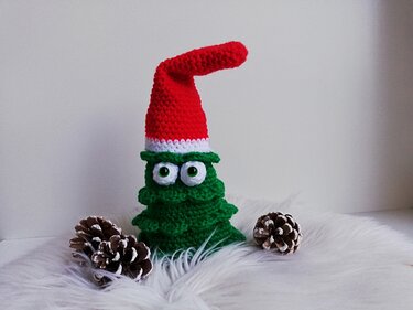 Gehäkelter grüner Amigurumi-Weihnachtsbaum mit großen Kulleraugen und roter Zipfelmütze auf weißem Fell, daneben Tannenzapfen