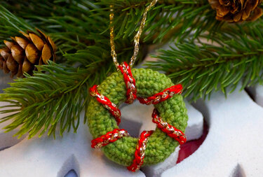 Gehäkelter grüner Mini-Kranz mit rotem und goldenem Band, umgeben von Tannengrün und Zapfen.
