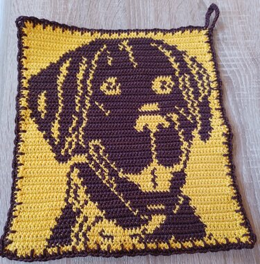Quadratischer gehäkelter Topflappen mit Labrador‑Kopf in Braun auf gelbem Hintergrund und Aufhängeschlaufe.