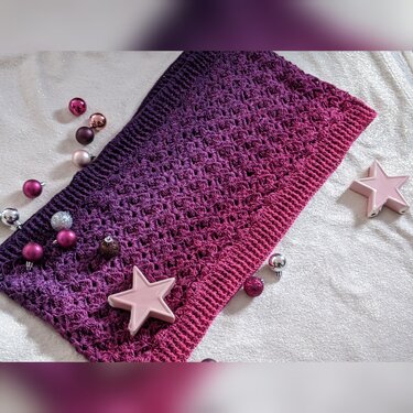Gehäkelter Schal in Violett‑Pink mit offenem Muster, dekoriert mit kleinen glänzenden Weihnachtskugeln und rosa Sternen auf hellem Stoff.