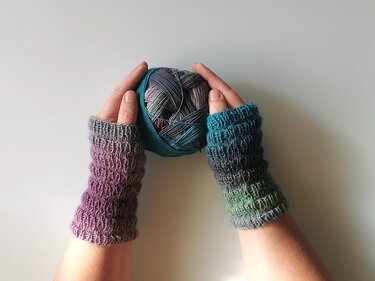 Hände mit fingerlosen Armstulpen aus Verlaufsgarn (Lila bis Grün), Maschenstruktur gut sichtbar, halten ein mehrfarbiges Garnknäuel
