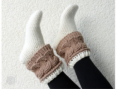 Beine in schwarzen Leggings mit cremeweißen Häkelsocken und beigebraunen, zopfartigen Stulpen vor heller, strukturierter Wand.