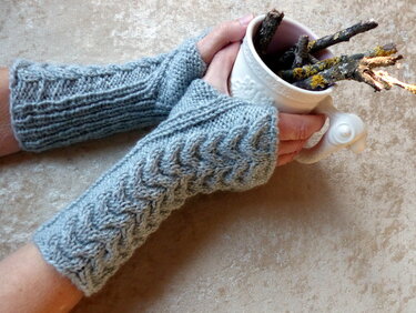Grau gestrickte, fingerlose Stulpen mit Zopfmuster an zwei Händen, die eine Tasse halten