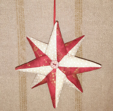 Gesteppter achtzackiger Stoffstern in Rot und Creme mit goldfarbenem Notenmuster, Knopf in der Mitte, hängend