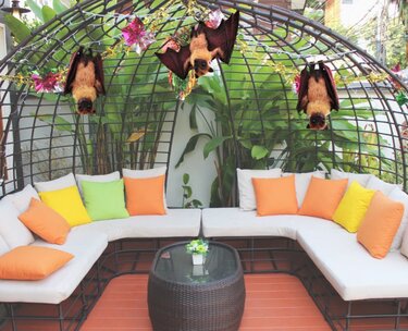 Three crochet bat amigurumi hanging upside down from a decorated metal trellis above an outdoor lounge with colorful cushions