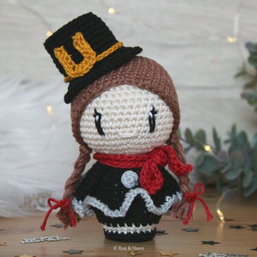 Gehäkelte Amigurumi-Puppe mit Zylinderhut, rotem Schal und geflochtenen Zöpfen vor Lichterkette