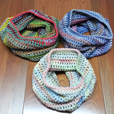 Drei gehäkelte Loop‑Schals in verschiedenen bunten Farbverläufen auf Holzfußboden