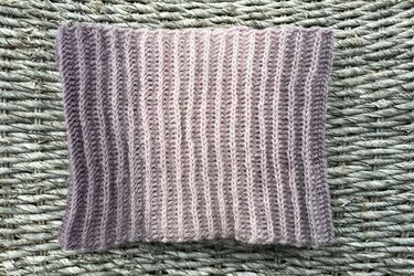 Gehäkelter Loop‑Schal mit vertikalem Rippenmuster und Lila‑Rosa‑Farbverlauf auf geflochtenem Untergrund