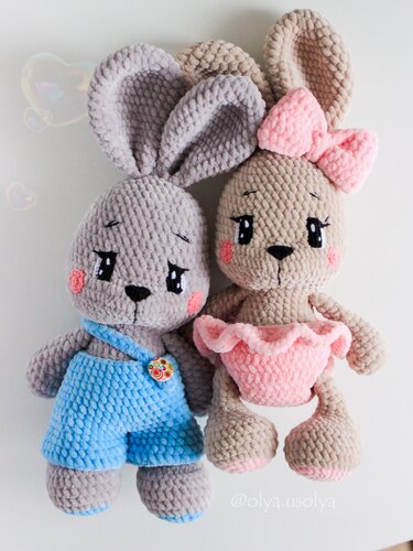 Two plush crochet bunny dolls, grey in blue overalls and beige with pink bow and ruffled skirt