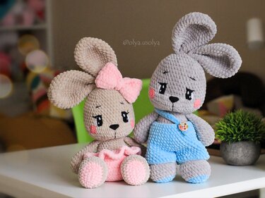 Two crocheted amigurumi bunnies, one beige with a pink bow and skirt and one grey in blue overalls, seated on a white surface.