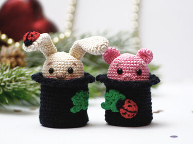 Zwei gehäkelte Mini-Amigurumi (Hase und Schwein) in schwarzen Töpfchen. Der Hase trägt einen Marienkäfer auf dem Ohr. Die Töpfchen sind mit Kleeblatt und einer roten Blumenapplikation versehen.