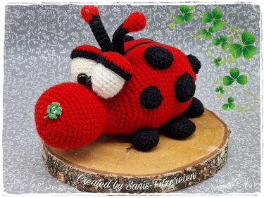 Roter gehäkelter Amigurumi-Marienkäfer mit schwarzen Punkten, weißen Augen und grünem Kleeblatt-Button auf einer Baumscheibe.