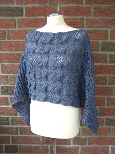 Blau-grauer gestrickter kurzer Poncho mit Ajour- und wellenartigem Muster auf Mannequin vor Ziegelwand