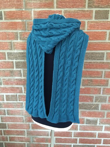 Petrolfarbener gestrickter Kapuzenschal mit Zopfmuster auf einer Schneiderpuppe vor Backsteinwand.