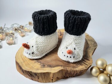 Handgestrickte Babyschuhe mit Schneemann-Gesicht und orangefarbener Nase auf einer Holzscheibe.