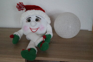 Gehäkelte Weihnachtsfigur mit roter Zipfelmütze und grünen Stiefeln neben glitzernder weißer Kugel