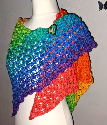 Gehäkeltes Dreieckstuch mit Regenbogen-Farbverlauf, auf weißer Schneiderpuppe drapiert und mit Herz‑Brosche befestigt