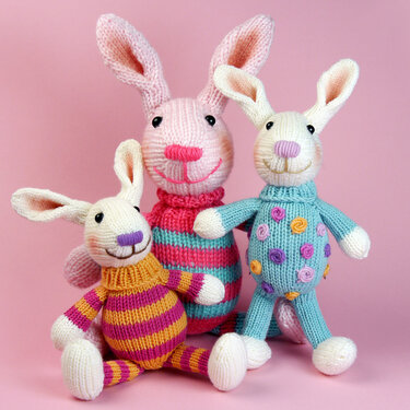 Three pastel knitted amigurumi bunnies wearing striped and dotted sweaters on a pink background