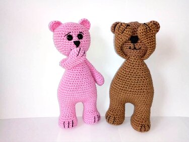 Zwei gehäkelte Amigurumi-Teddys (rosa und braun); rosa hält Hand vor Mund, braun bedeckt die Augen; weißer Hintergrund.
