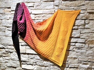 Gestricktes dreieckiges Tuch mit Farbverlauf von Violett über Orange zu Gelb, mit Wabenstruktur und Motivfeldern, hängend an einer hellen Steinwand