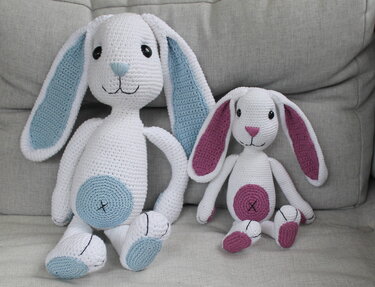 Zwei gehäkelte Amigurumi-Hasen in Weiß mit langen Schlappohren, groß und klein, mit blauen und magentafarbenen Akzenten auf einem grauen Sofa.