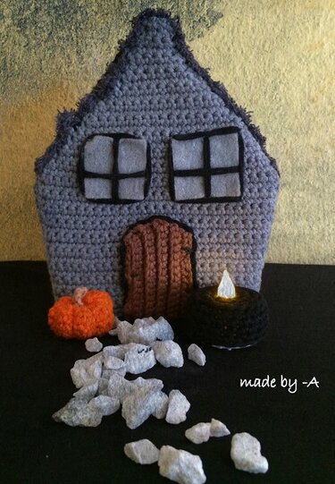 Gehäkeltes Spukhaus in Grau mit Fenster- und Türdetails, Amigurumi‑Kürbis, LED‑Teelicht und weißen Dekosteinen